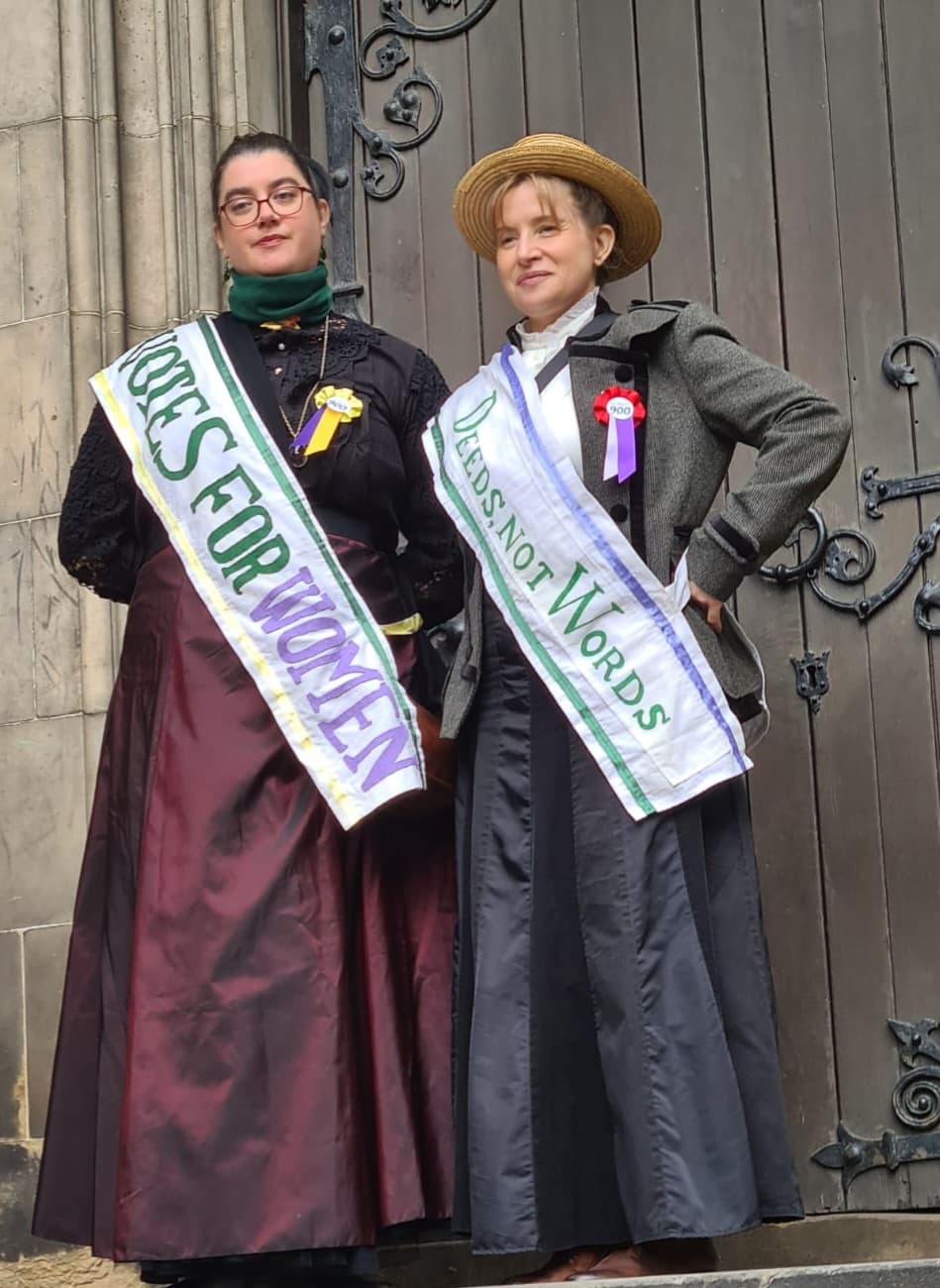 Two women dressed as suffragettes wearing sashes saying Votes for Women