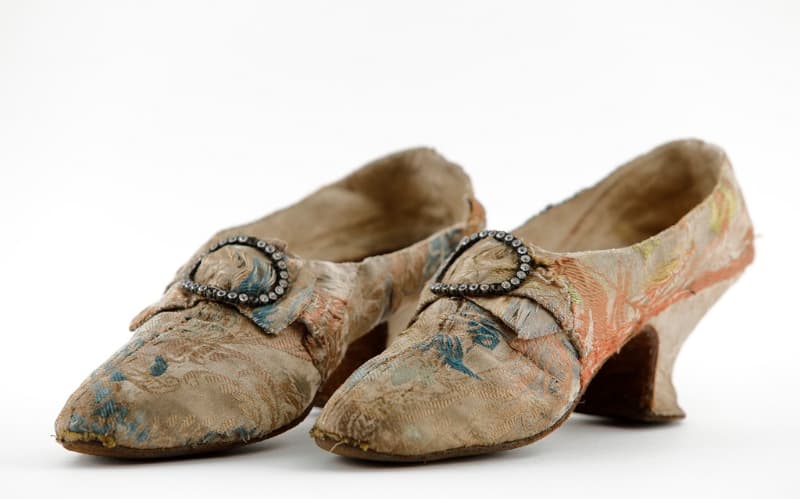 A pair of high heel shoes dating back to the 1750s featuring a buckle and faded cream, blue and orange fabric