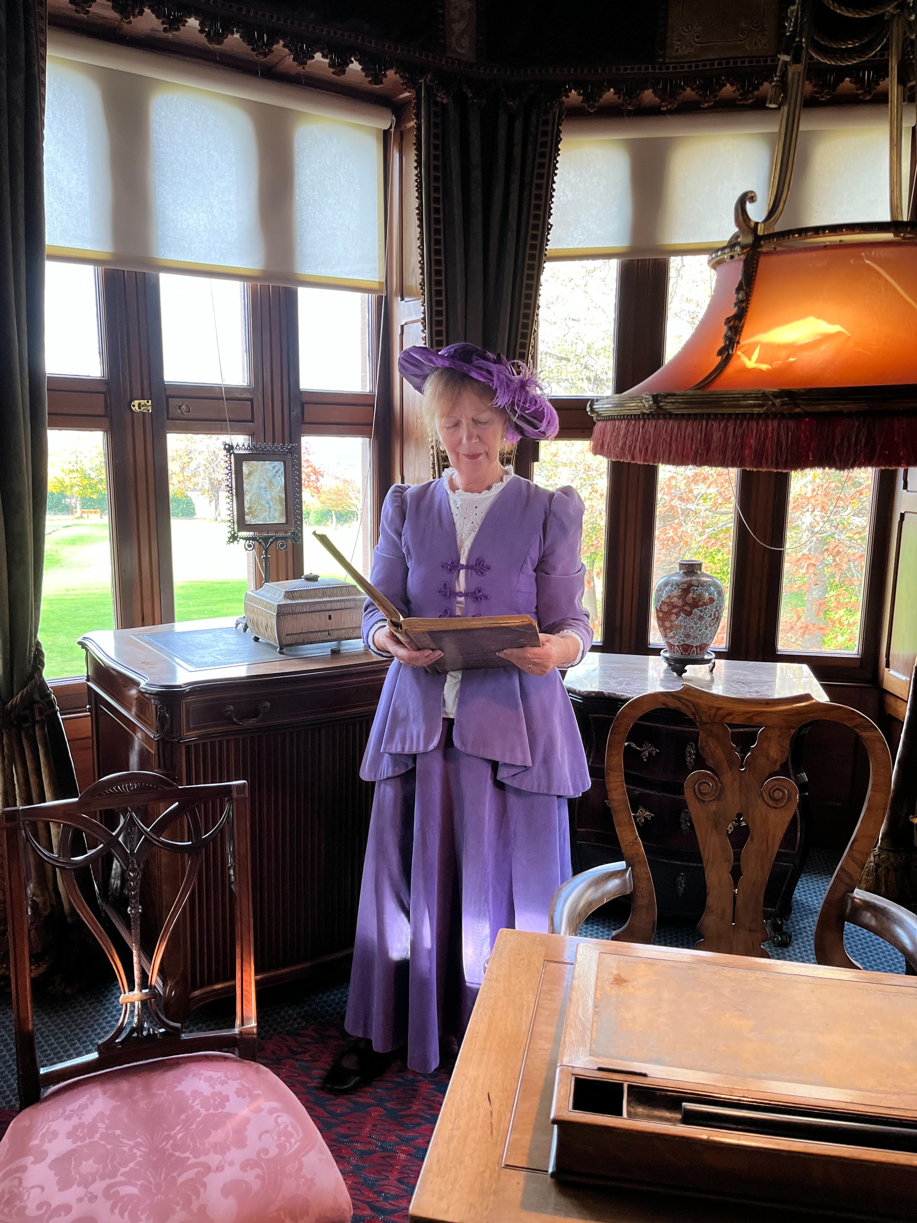 A women in Edwardian costume stand beside a window in a grand room 