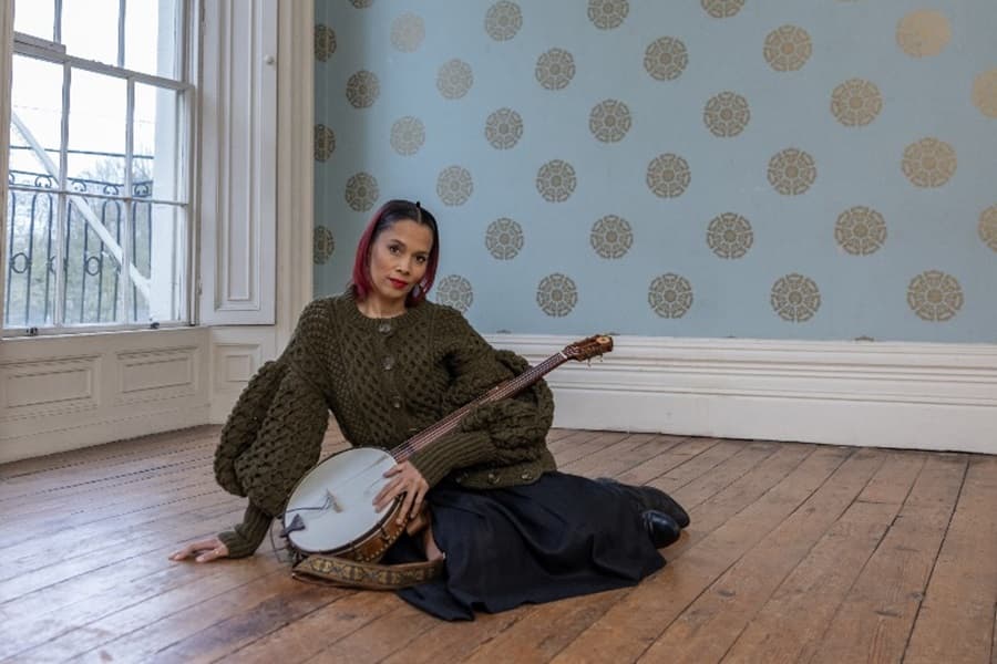 Rhiannon Giddens sitting on the floor holding her banjo on her lap.