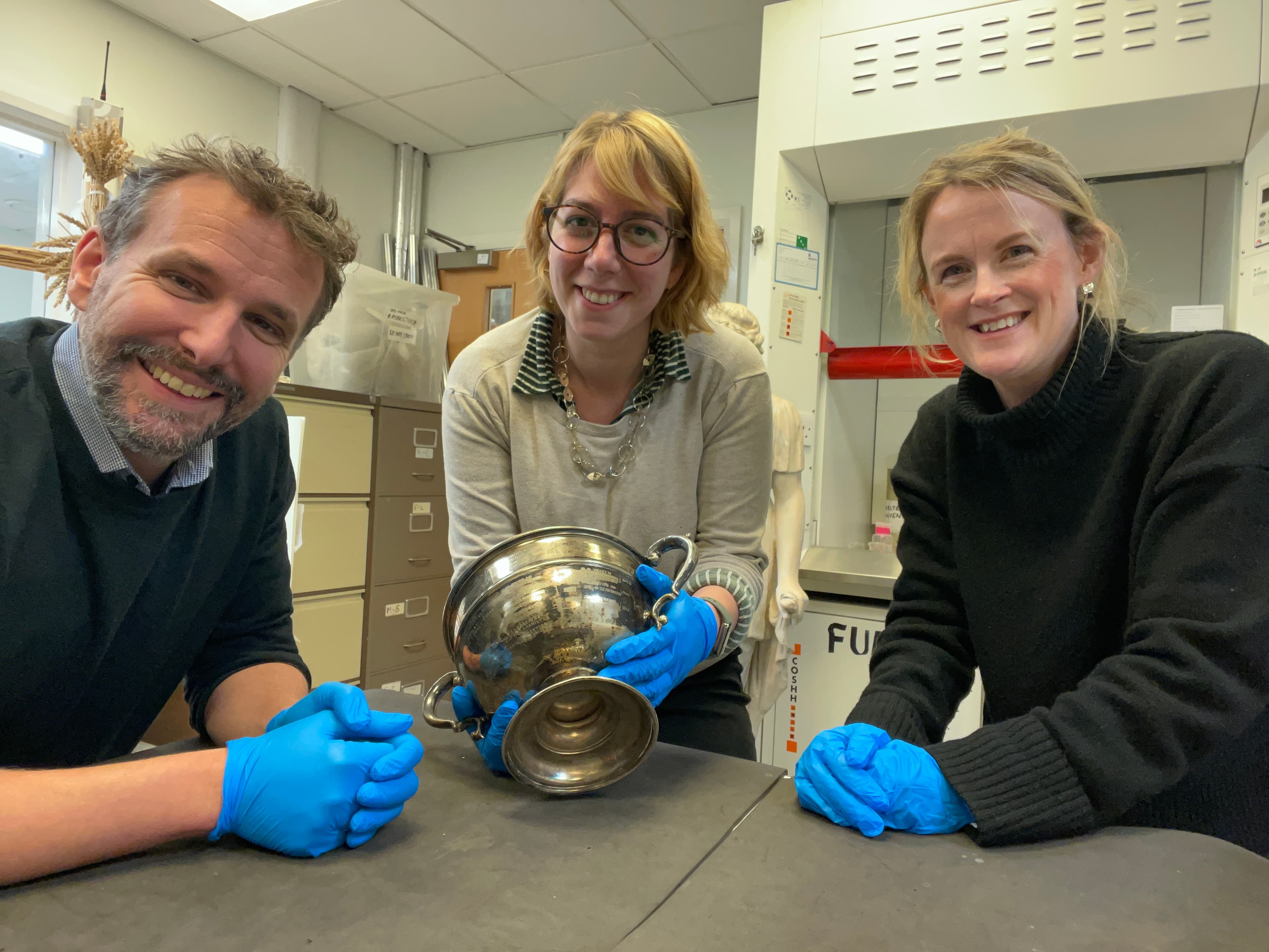 Three smiling people. One person holds a vintage silver pot. 