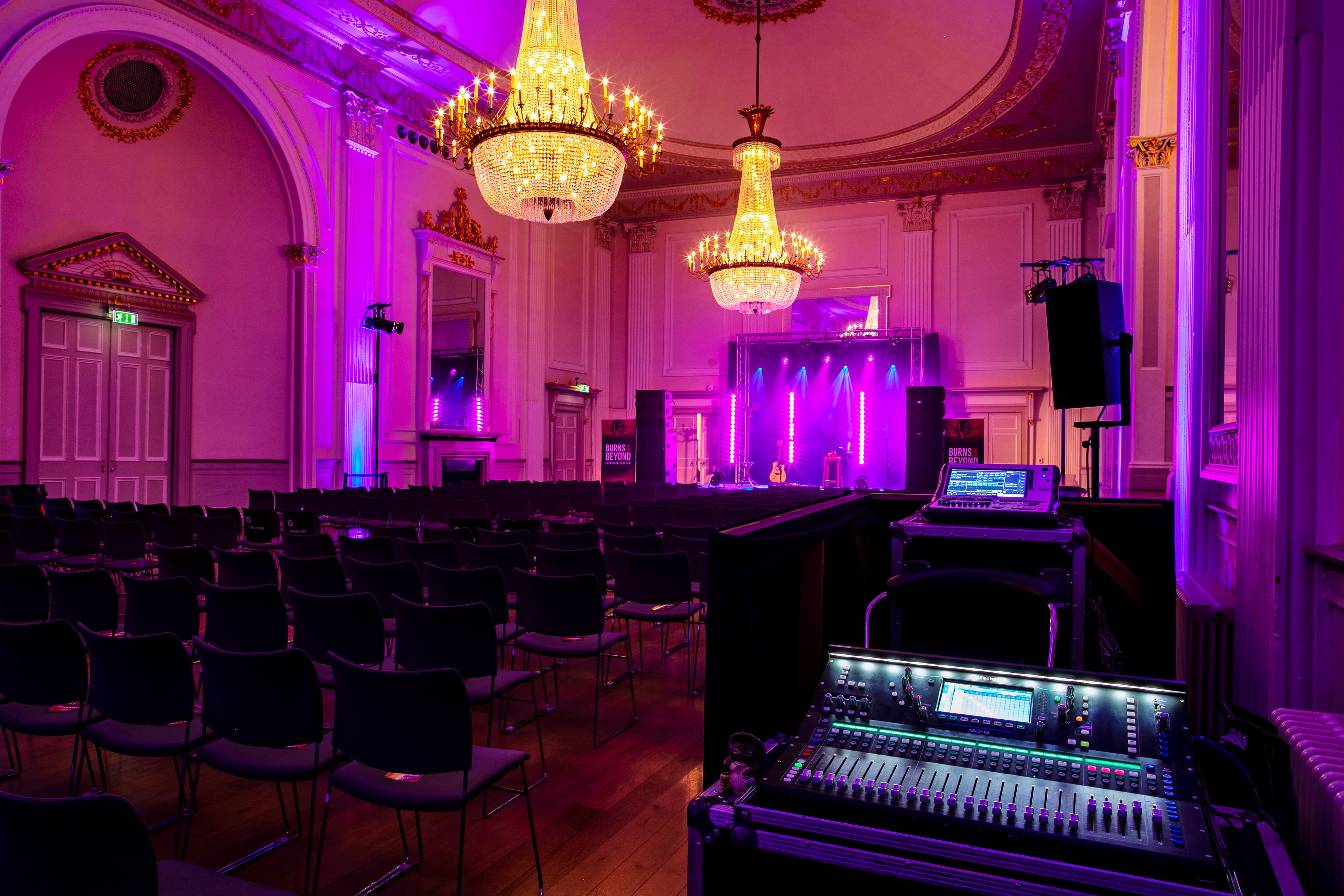 A spacious room illuminated with purple lighting, featuring a stage at the front for performances or presentations. Control deck in the foreground.
