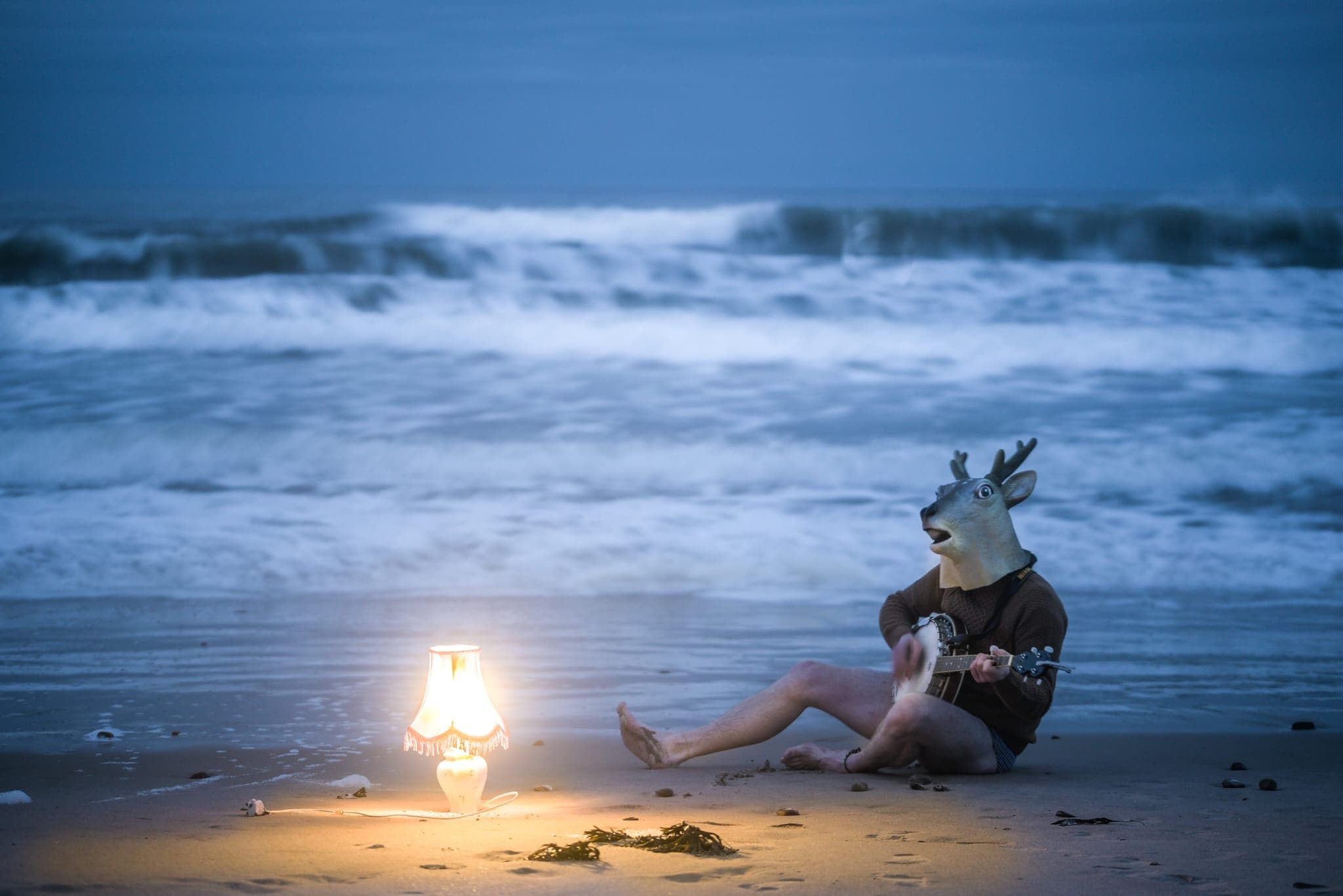 A man sat on the beach wearing a fake deer head and playing banjo next to a lit lamp 