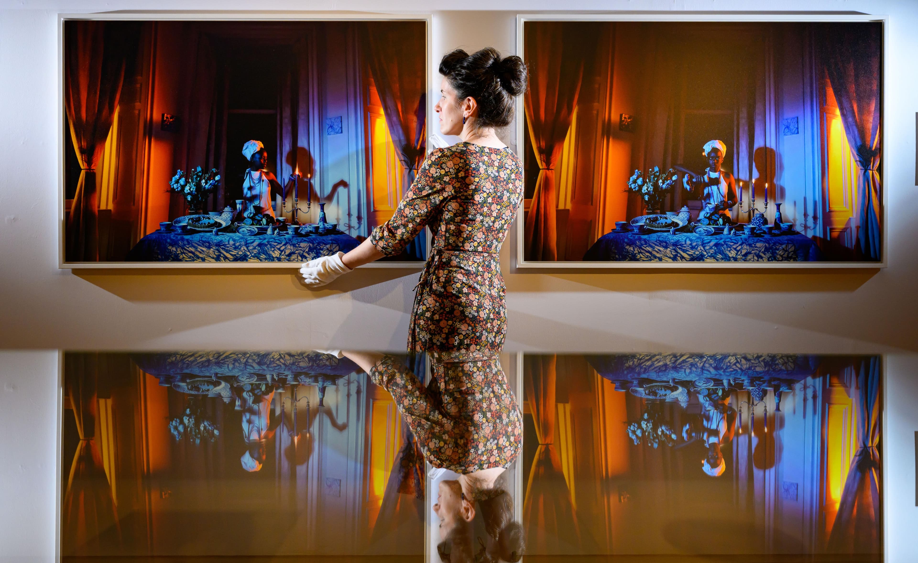A curator in white gloves adjusts the position of an artwork while standing between two pieces on display on a wall.