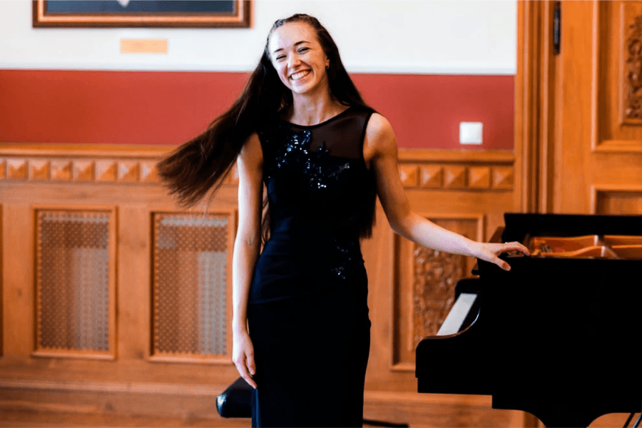 Pianist, Catriona MacKenzie, standing with their left hand placed on the piano smiling