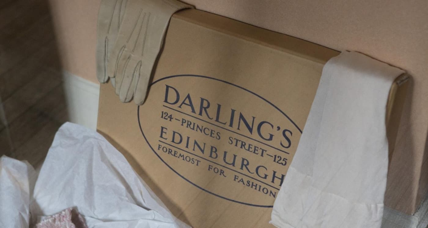A brown box from the old department store, Darling's which was once located on Princes Street, Edinburgh. The box features text "Darling's 124 - Princes Street - 125 Edinburgh Foremost For Fashion" A vintage pair of white gloves and stockings are draped over the box.