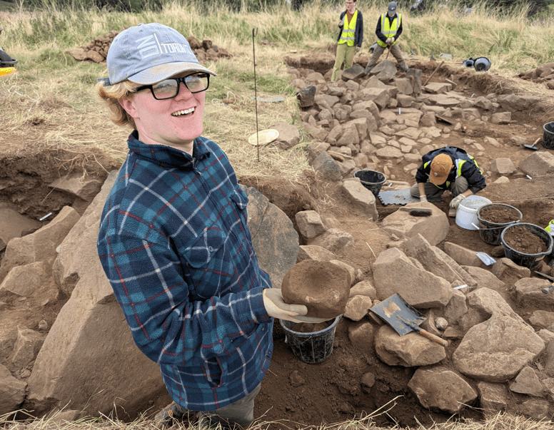 Male archaeologist smiling and holding archaeological findings while stood in a hole in the ground. Men in hi-vis vests are stood behind him also working.
