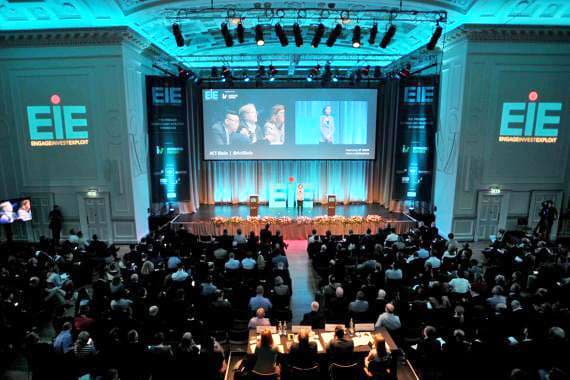 A large crowd focuses on a presentation taking place on a stage in a conference setting.