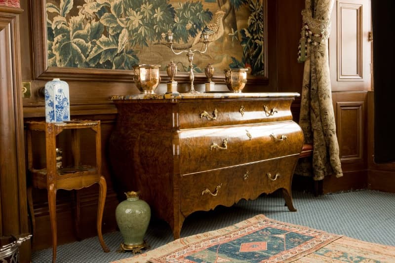 An Edwardian room in Lauriston Castle Edinburgh, featuring an Edwardian chest of drawers with vases and a candlestick on top. A jade green vase sits on the floor and a large tapestry in greens and browns is framed and displayed on the wall. 