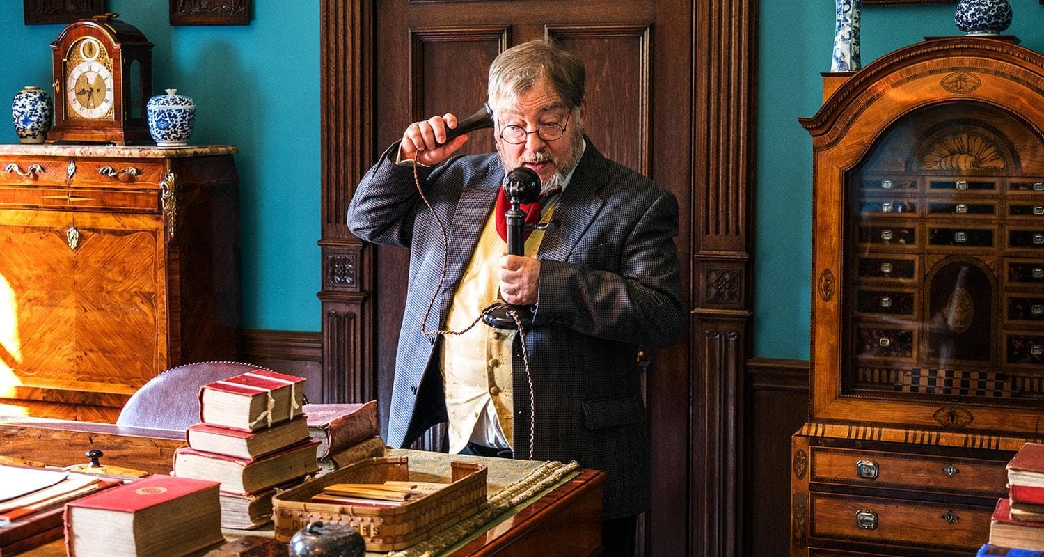 A man in an Edwardian suit and tie stands by a desk full of books and a vintage telephone in his hand, ready to speak.