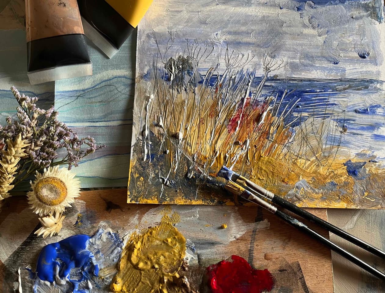 A painting depicting plants and the sea arranged on a wooden table with paint brushes and dried flowers.