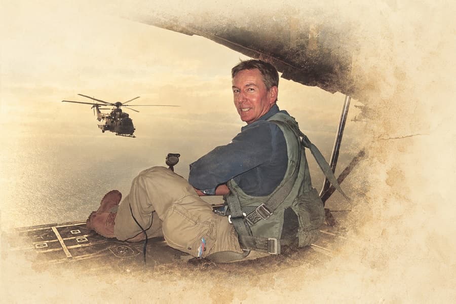 Image shows Frank Gardner sitting in lookout smiling with helicopter in background