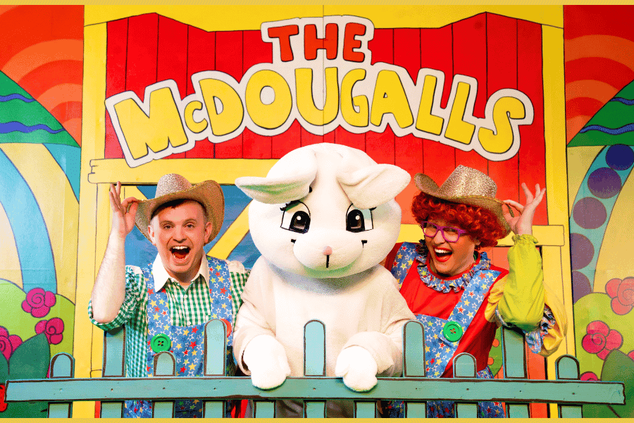 2 McDougalls Characters wearing colourfull clothing and cowboy hats standing behind farm fence with a large - human-sized white rabbit mascot between them