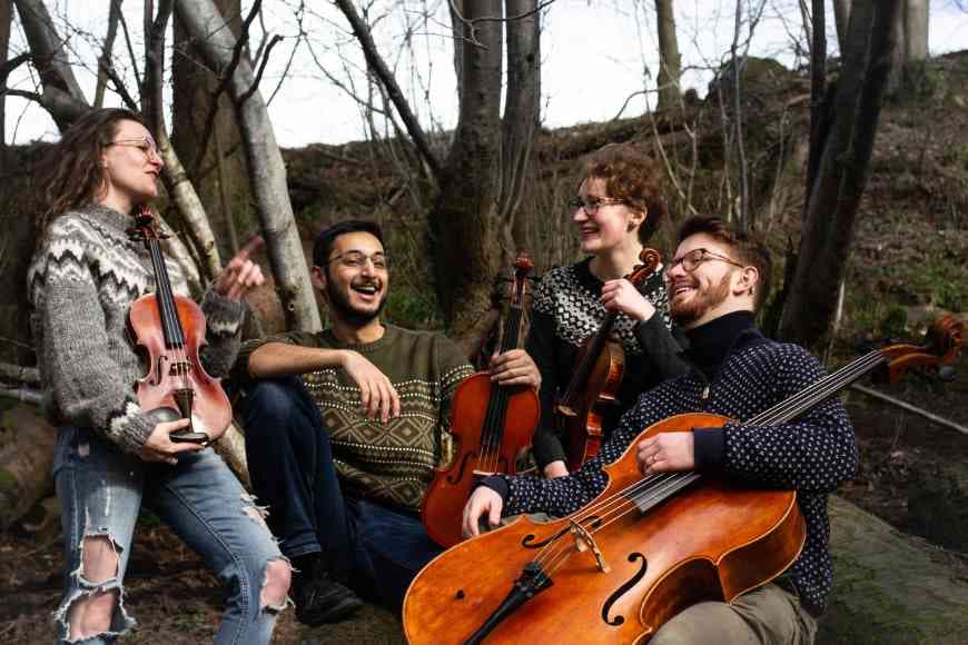 Photograph of members of Haver Quartet sitting in woodland laughing with instruments in hand.