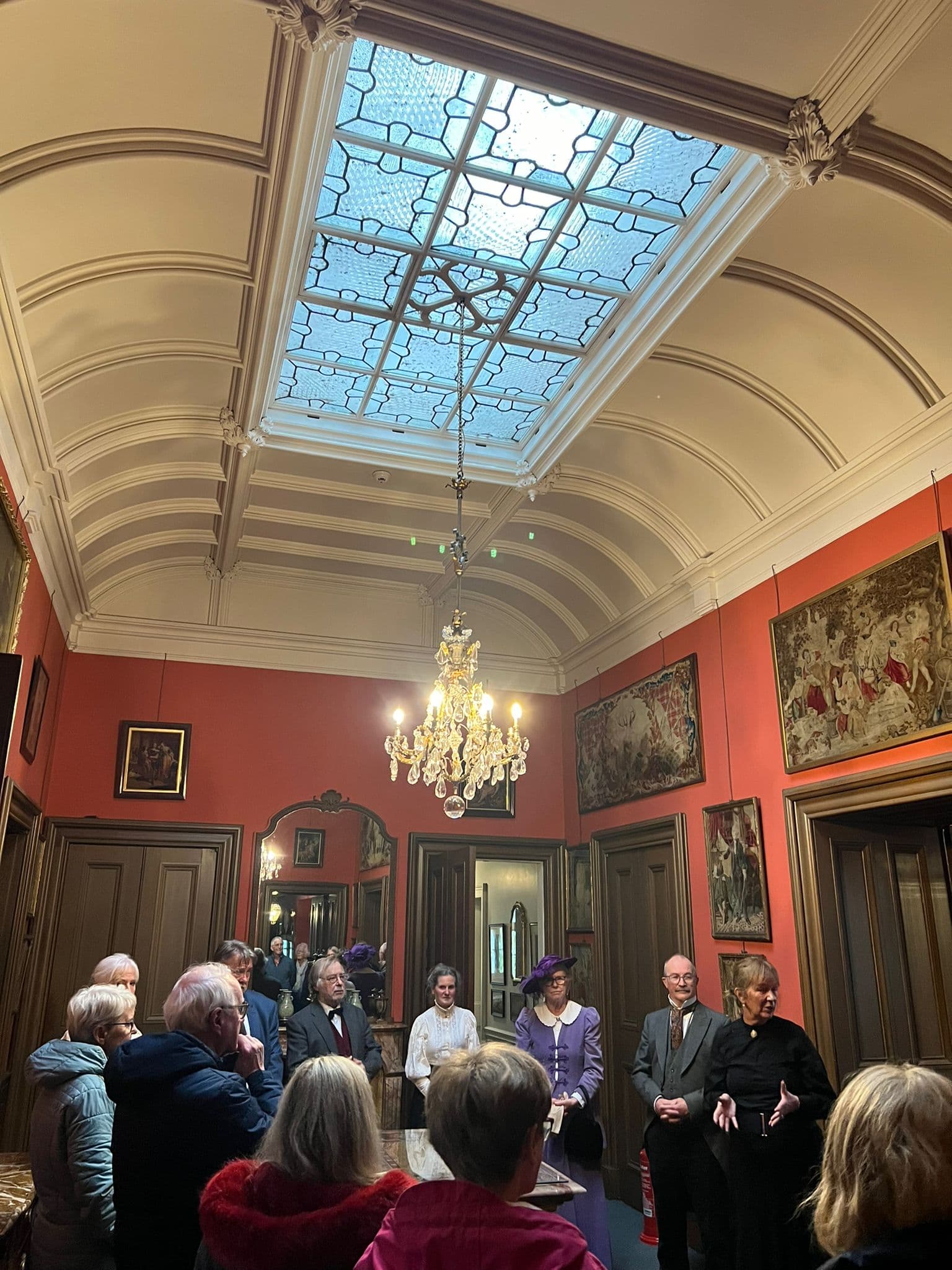 A group of visitors stand in the hallway at Lauriston Castle beneath a chandelier and enjoy a tour given by Edinburgh Living History group, dressed in Edwardian costume. 