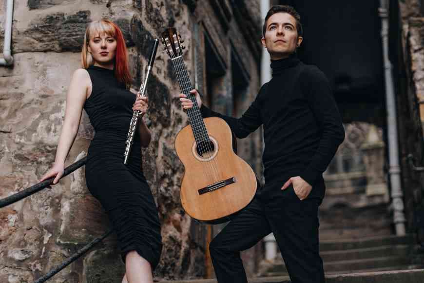 Photograph of Kerry Lazzaretti wearing sleeveless black dress holding her flute and Jacopo Lazzaretti wearing black poloneck jumper and black jeans holding his guitar standing on steps outside.