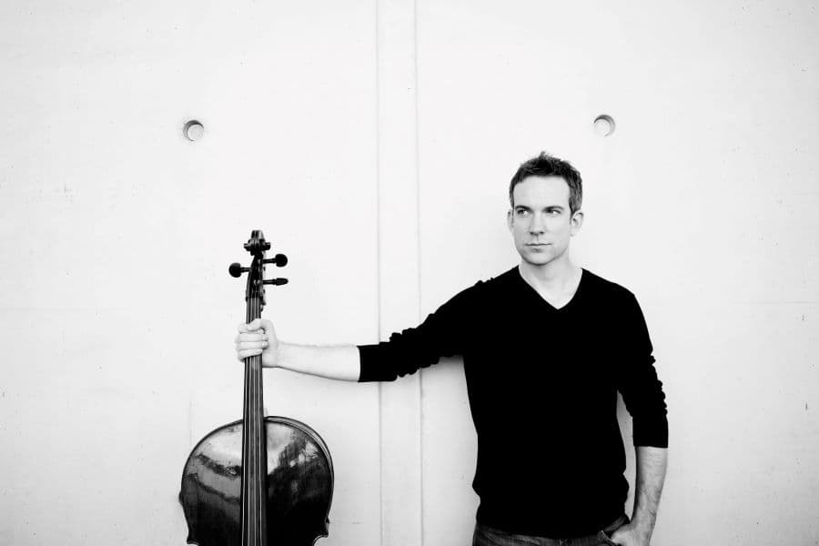 Black and white photograph of cellist, Johannes Moser standing holding his cello to the right of him