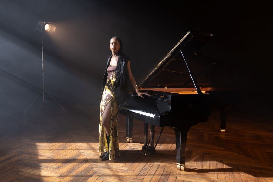 Jeneba Kanneh-Mason standing leaning against piano with her hands on the keyboard lid behind.
