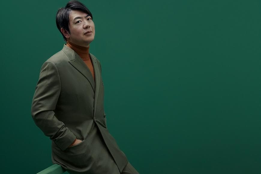 Lang Lang in a khaki colour suit and brown polo neck against a deep green background
