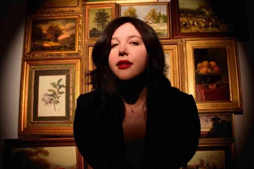Lucy Dacus standing in from of framed paintings.  The light is dimmed and her face is in spotlight.