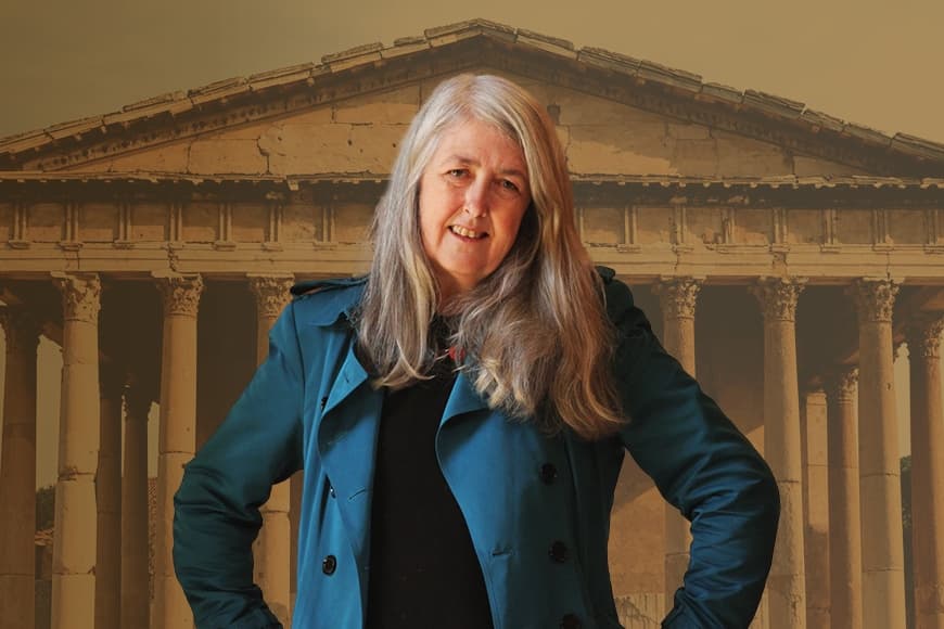 Mary Beard wearing a blue coat in from of a image of Roman temple