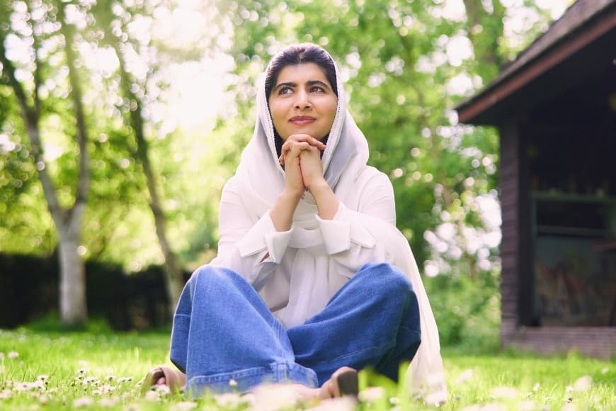 A photo of Malala Yousafzai sat cross legged on the grass with her hands clasped under her chin 