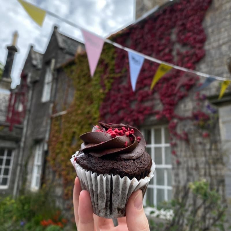 A hand holds a chocolate cupcake in front of Mimi's Little Bakehouse at Lauriston Castle in Edinburgh.