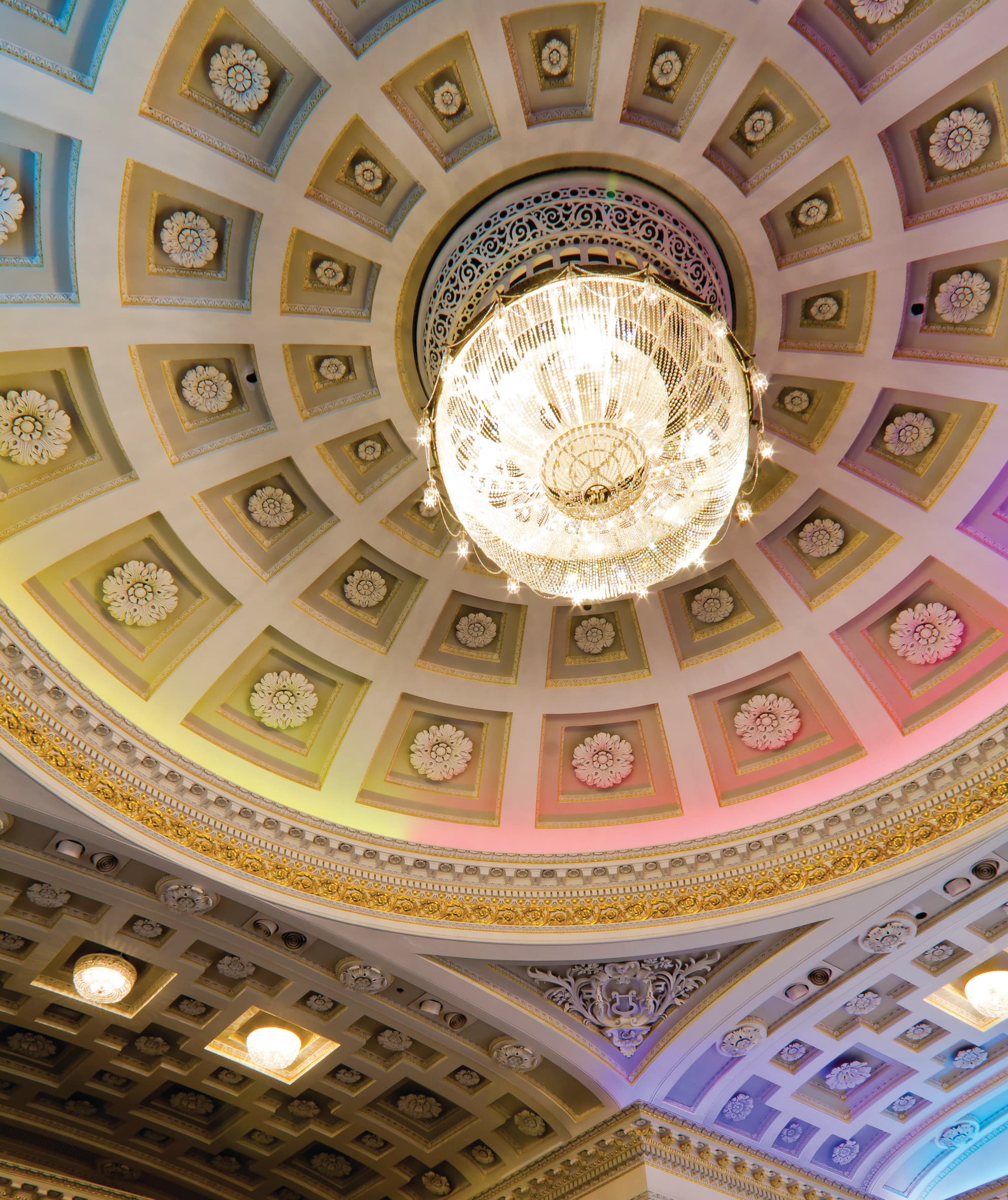 A ceiling featuring an ornate chandelier and architectural details, creating an elegant atmosphere. Lightening is positioned to show these details to the best ability.