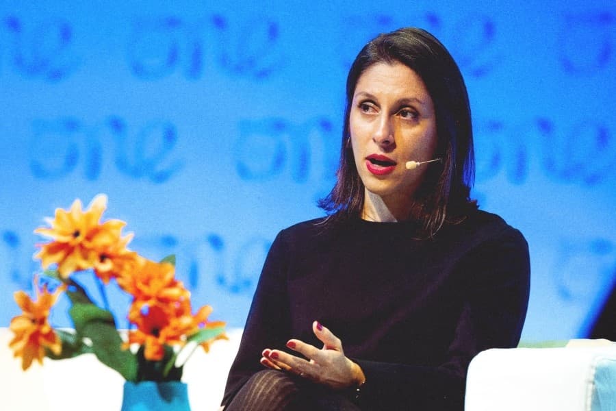 Nazanin Zaghari-Ratcliffe, dressed in black, with red lips and nails.  She is seated beside a blue vase with orange flowers.  