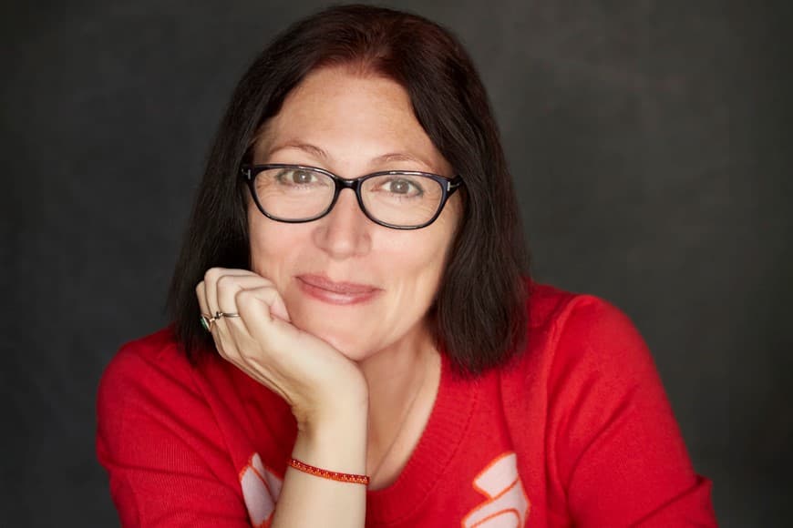 Photograph of Natalie Haynes with chin resting in right hand wearing a red jumper and smiling.