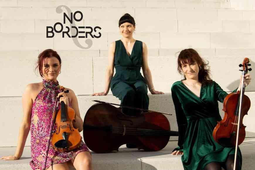 Musicians Frances Patterson (violin), Suzanne Godet (viola) and Joanna Stark (cello) who are No Borders Trio sitting, outside on a steps in the sunshine 