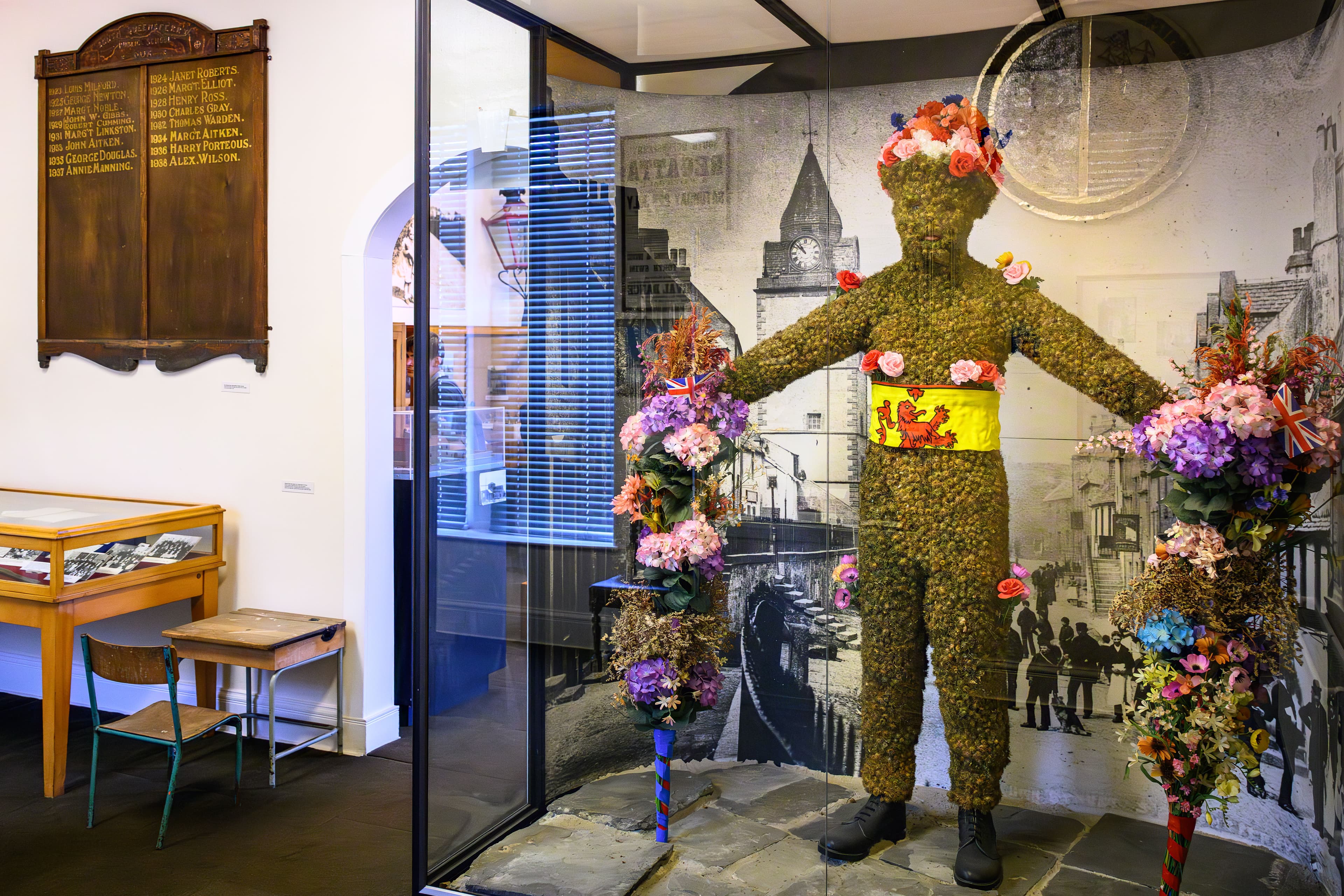 A model of a person in a suit made entirely of colorful burrs, standing confidently with a vibrant floral backdrop.