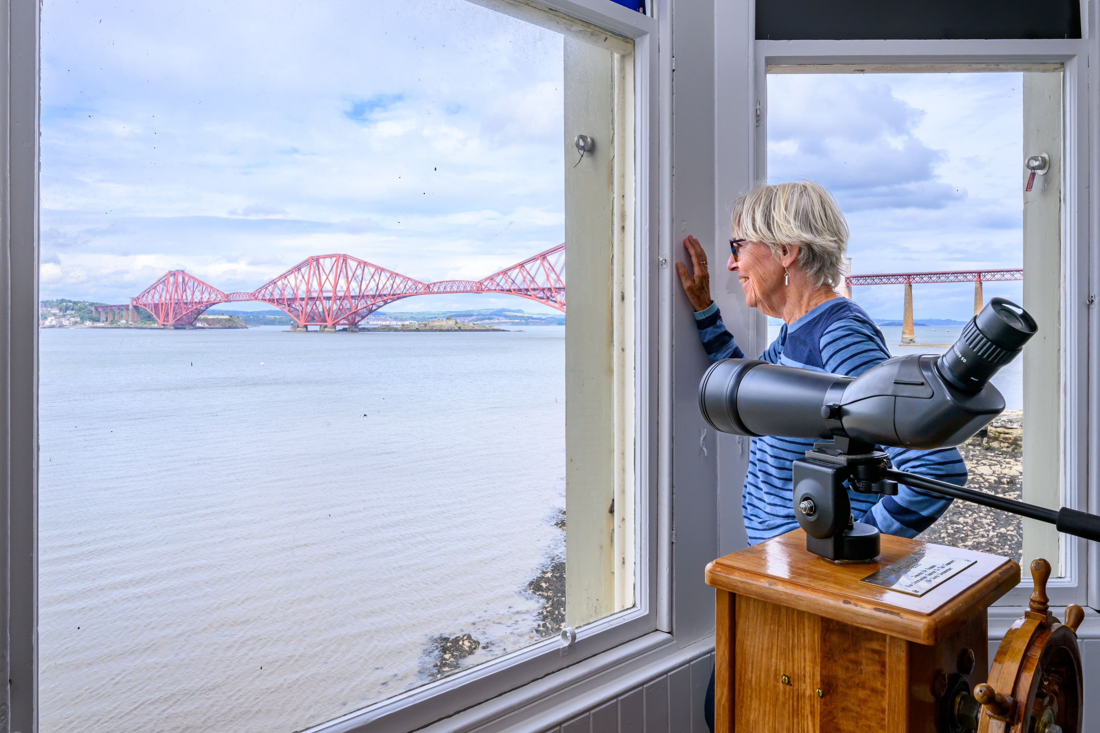 A woman gazes out a window in the museums telescope room, observing a bridge in the distance against a clear sky.