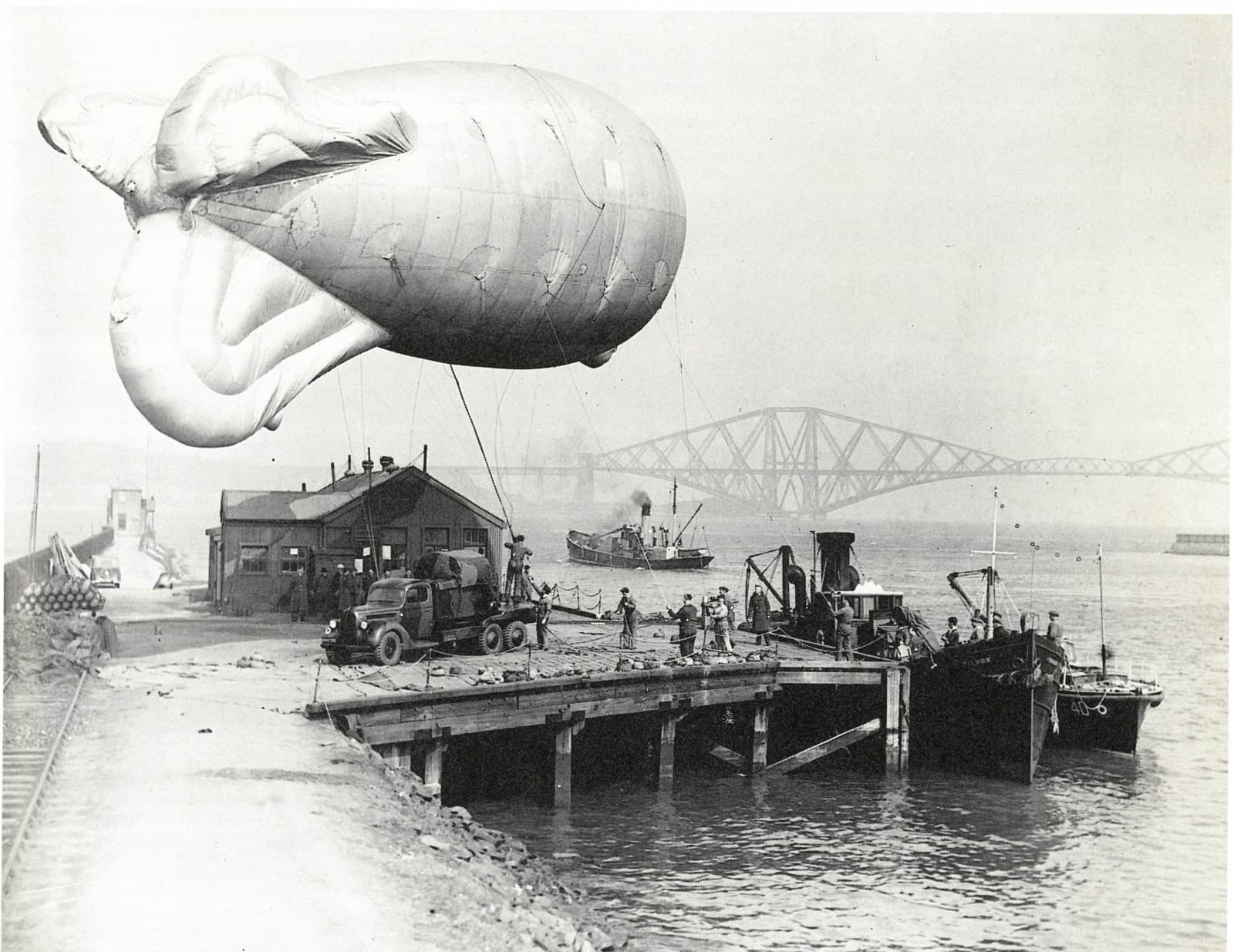 A photograph from the second world war featuring a zeppelin anchored near a dock on the River Forth with several vehicles, people, and boats in the background; the Forth Rail Bridge spans the river behind them.