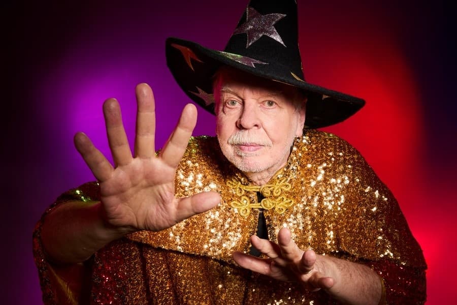 Rick Wakeman wearing pointy wizard's hat and gold cloak - arms outstretched like he is casting a spell.