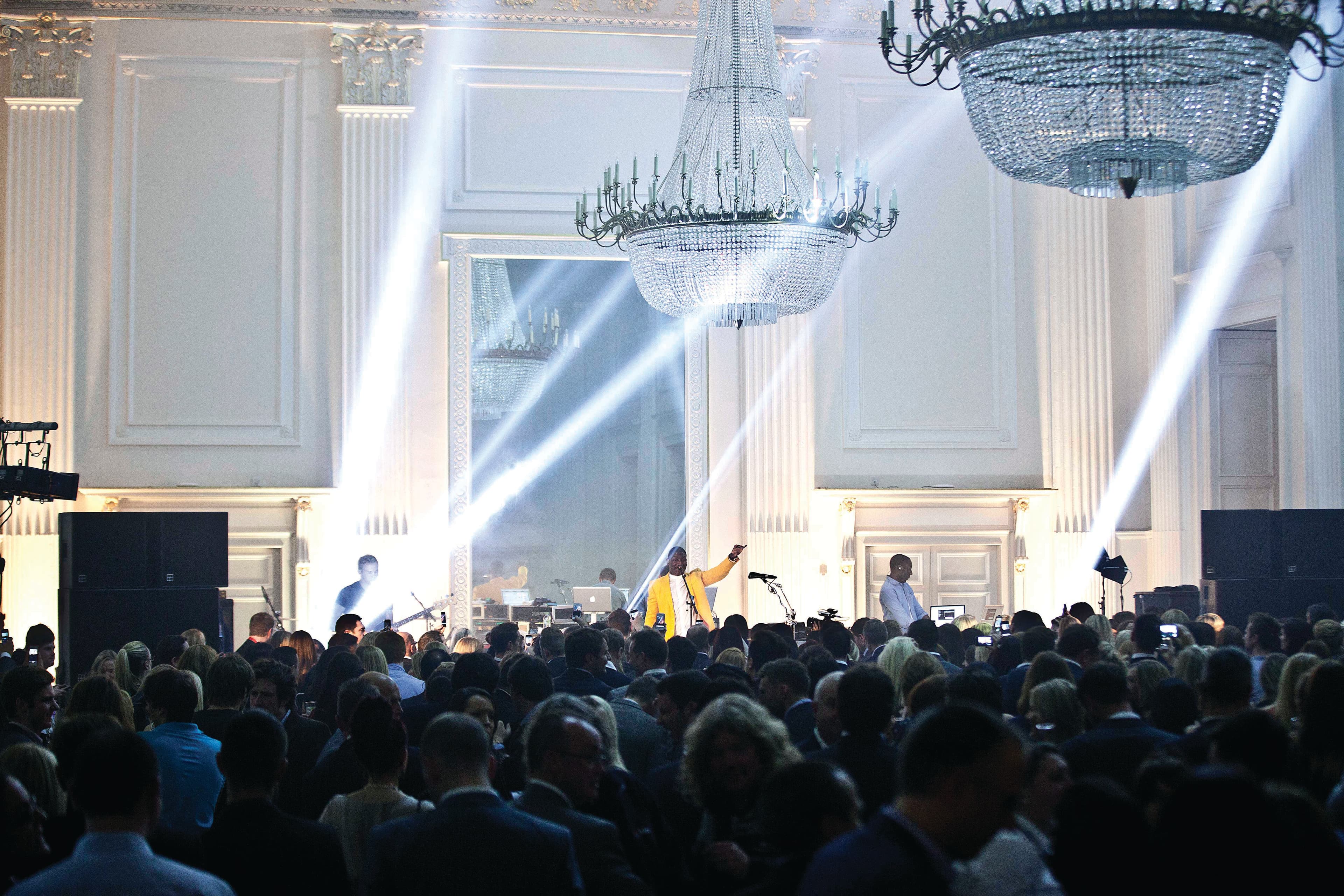 A bustling crowd fills a large room, with people interacting and moving about. The room has chandeliers and a large mirror. Spotlights light up the room from the floor.