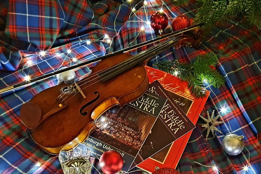 A fiddle lying on top of tartan alongside Scottish Fiddle Orchestra programmes surrounded by fairy lights