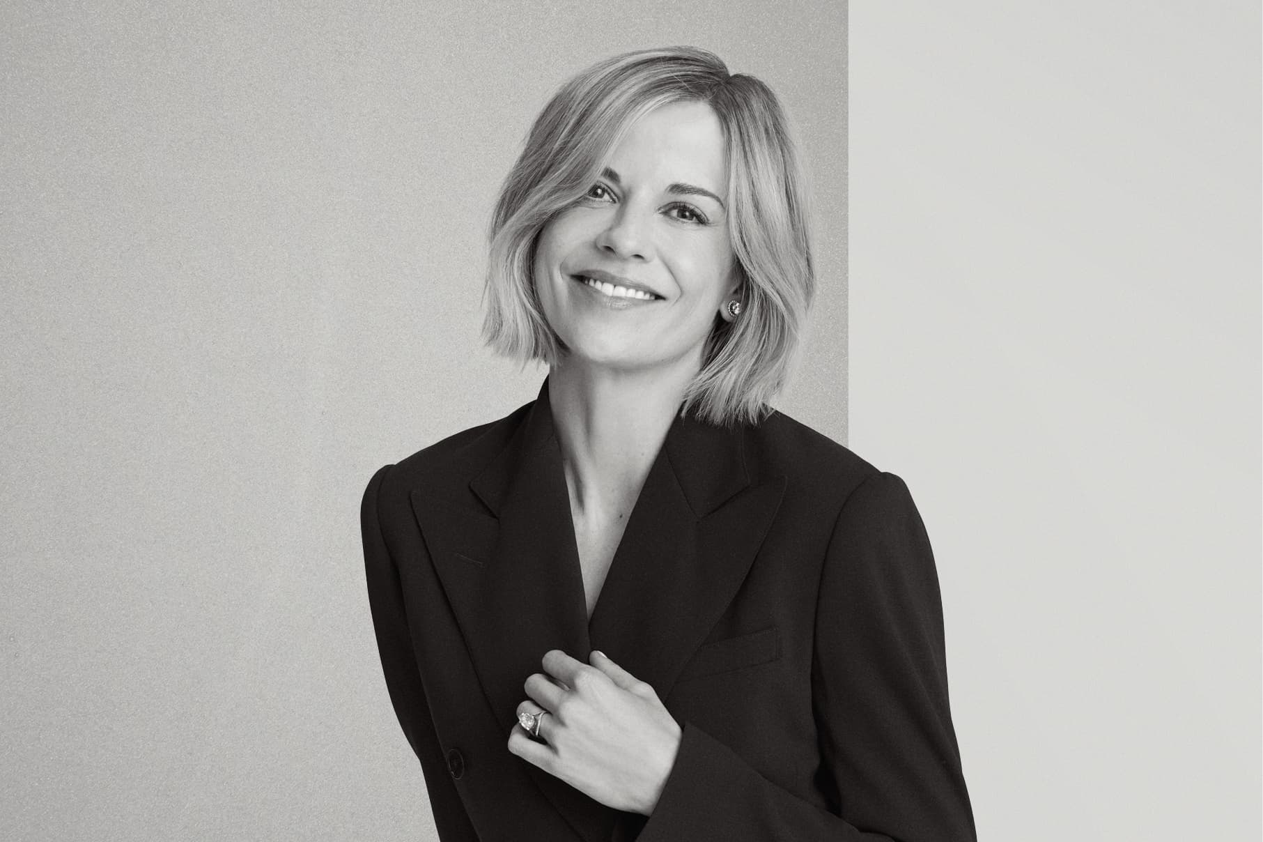 Black and White of Susie Wolff smiling at the camera with her left hand holding the lapel of the dark jacket she is wearing.