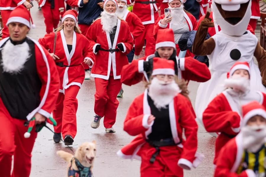 A crowd of people wearing Santa Claus costumes participate in a race, including a person in an Olaf snowman mascot suit and a dog running in the foreground.