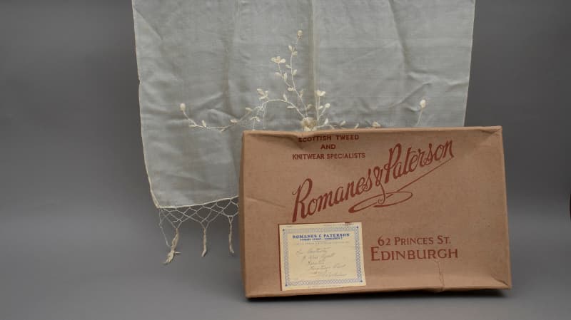 A vintage delicate white silk scarf with a fringe and embroidered flowers and leaves is on display next to a box from an old Edinburgh shop with the following words featured on the front "Romanes & Paterson, Scottish tweed and knitwear specialists. 62 Princes Street Edinburgh"