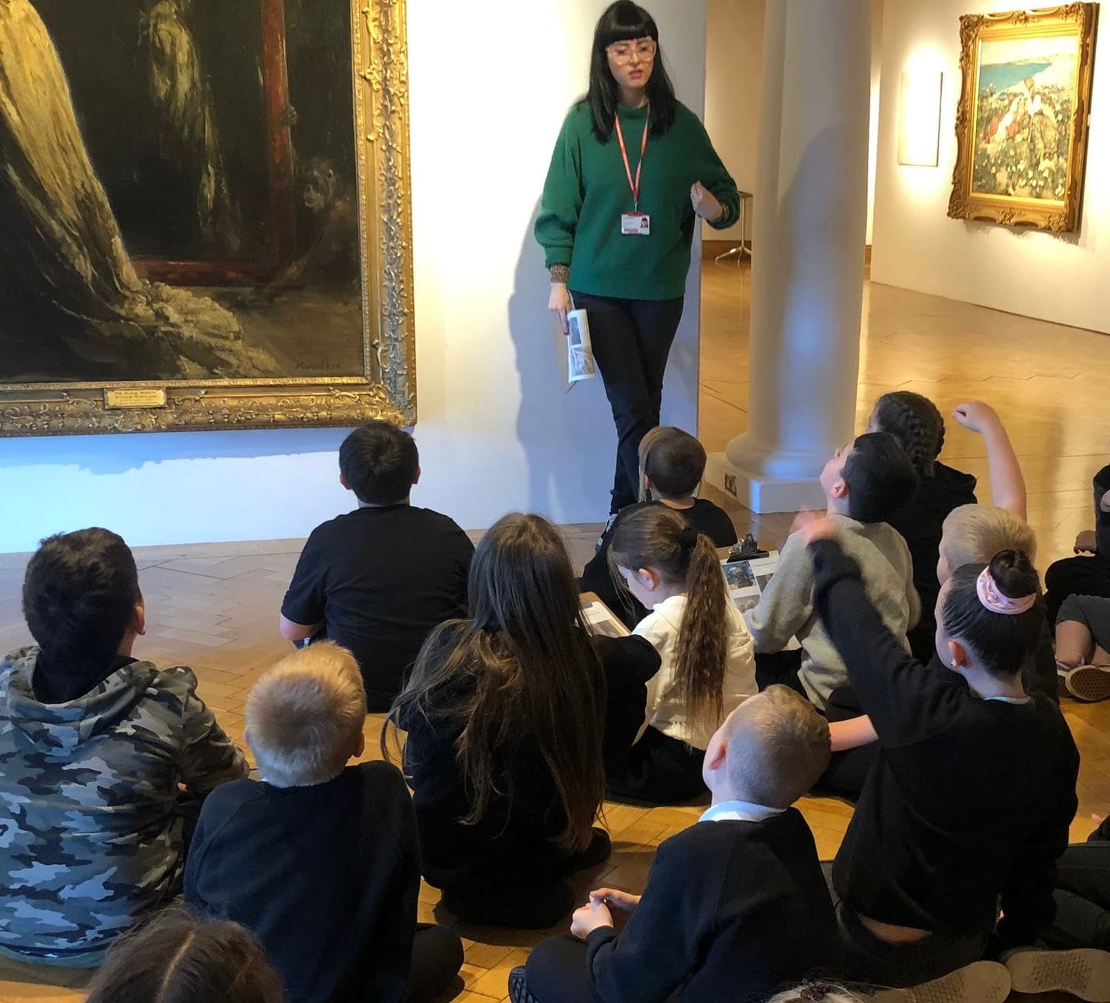 A teacher stands in front of a large painting in the City Art Centre in Edinburgh and addresses a group of school children sitting on the floor.