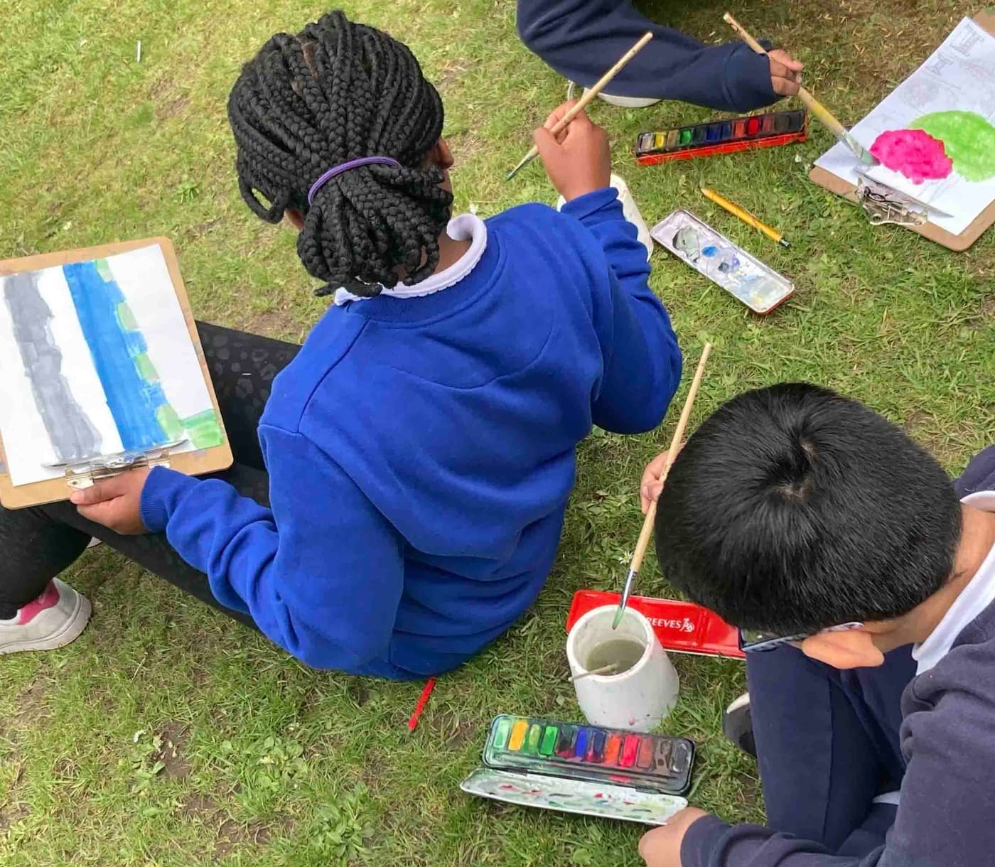 Two school children sitting on the grass, engaged in painting with brushes and paper around them