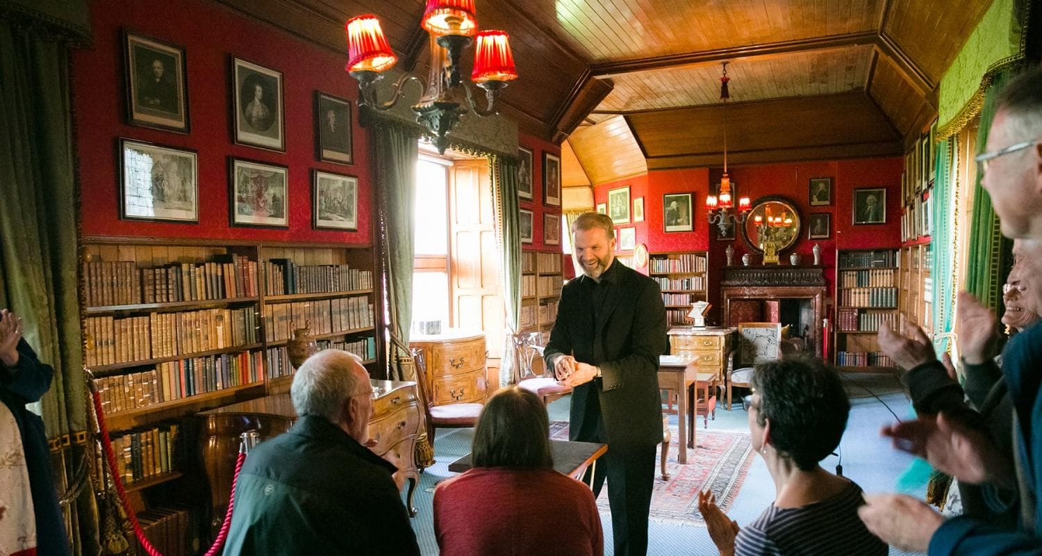 A magician in a black suit stands before a group of people in the library at Edinburgh's Lauriston Castle with a pack of cards in his hands. The library is full of vintage bookshelves, books and framed artwork on the walls.