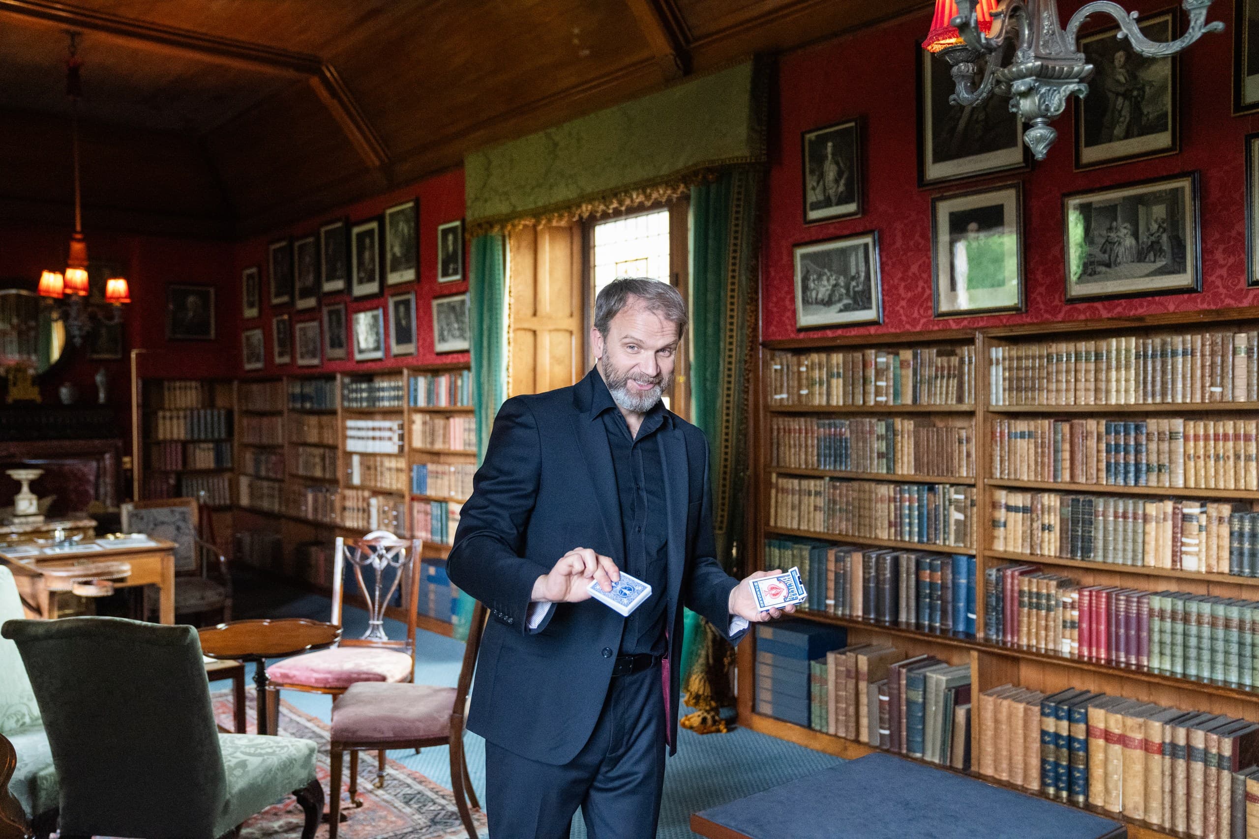 A magician holds a deck of cards and stands in the middle of the library of Edinburgh's Lauriston Castle. Antique bookcases are filled with books.  