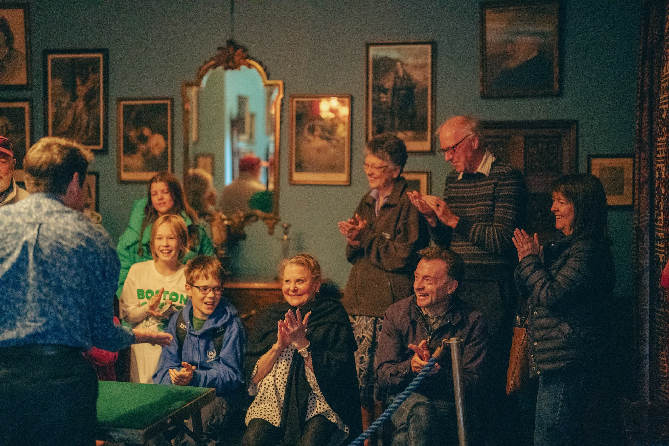 A group of people applaud a magician following his magic trick. The people are in a grand room in Edinburgh's Lauriston Castle surrounded by antique paintings and a gilt mirror on the wall.