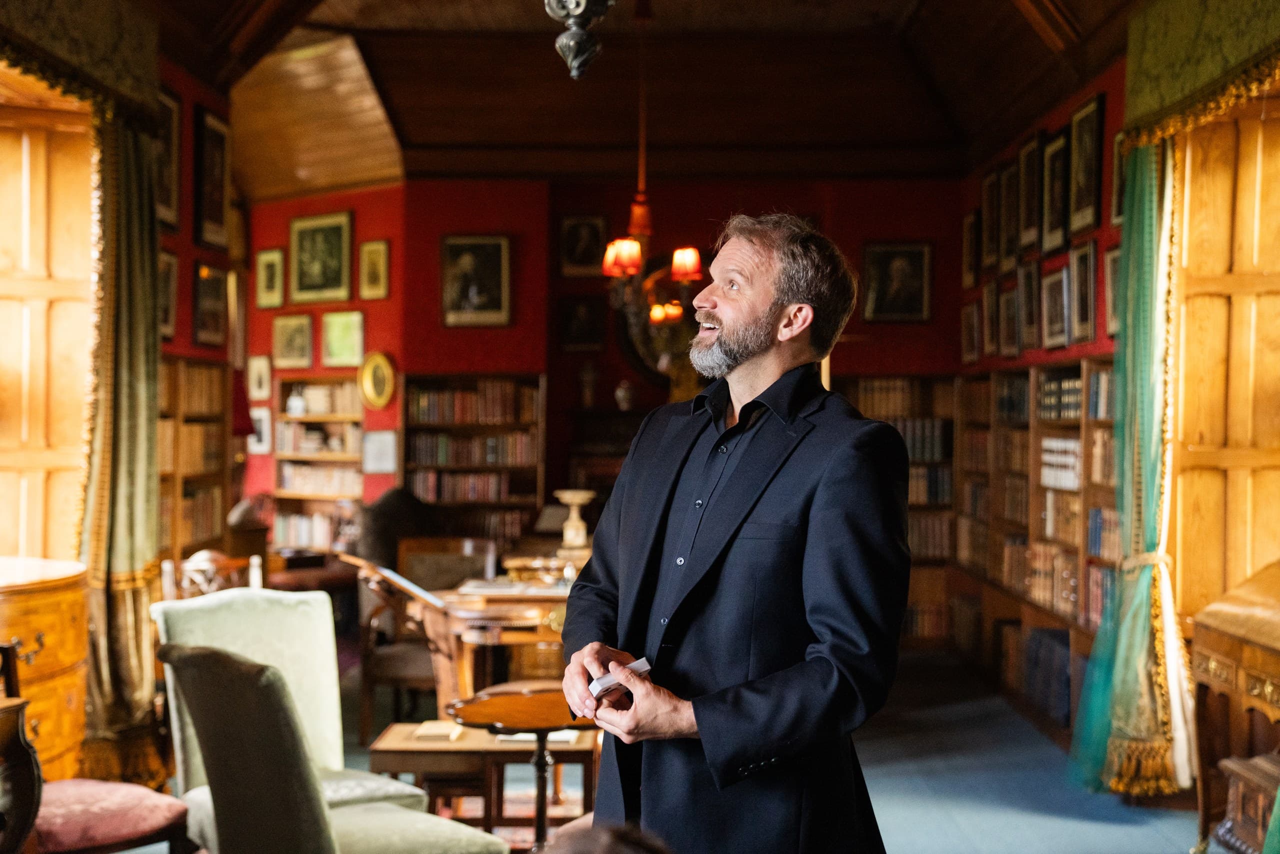 A man in a black suit stands in an Edwardian library at Edinburgh's Lauriston Castle. The room is full of vintage book shelves, book and framed artwork and photographs on the wall.