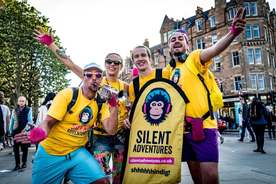 A cheerful group in bright yellow t-shirts and backpacks pose on a city street, one holding a sign, and one with a trumpet.