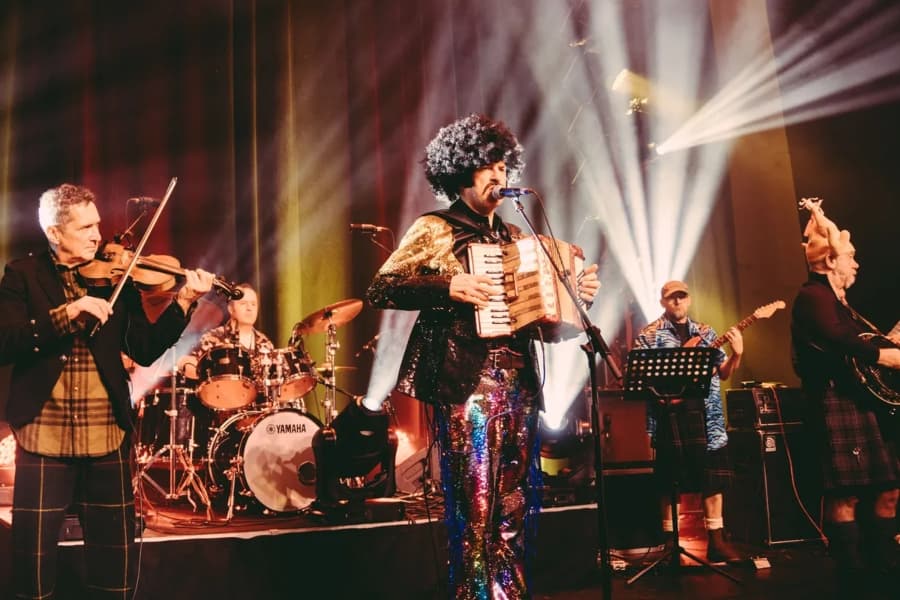 A photo of a band in funky 60s-70s outfits, frontman wearing a curly afro wig, playing traditional ceilidh instruments