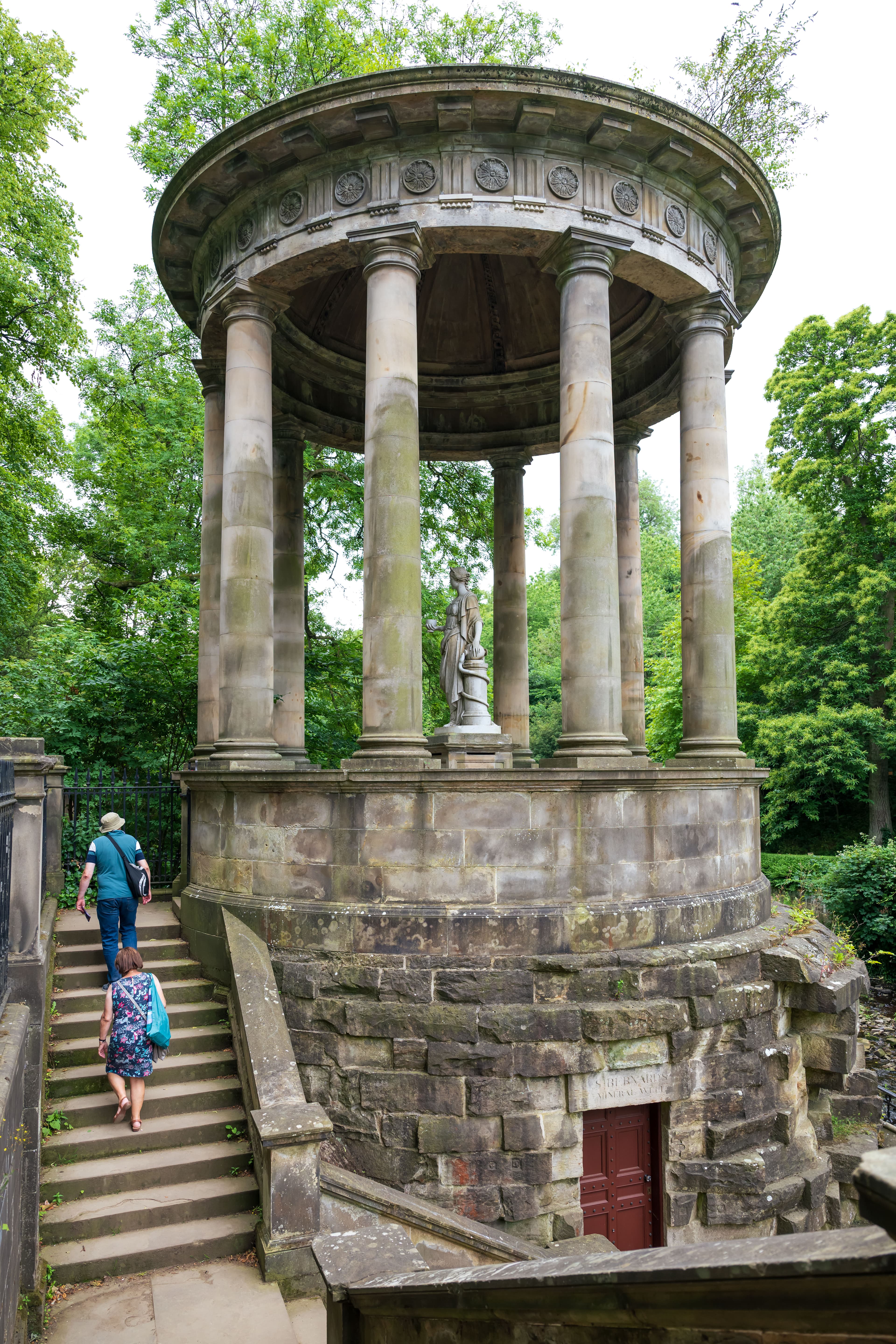 A stone monument surrounded by greenery, featuring a domed roof and open sides for a scenic outdoor view. Visitors are climbing steps to see more closely.