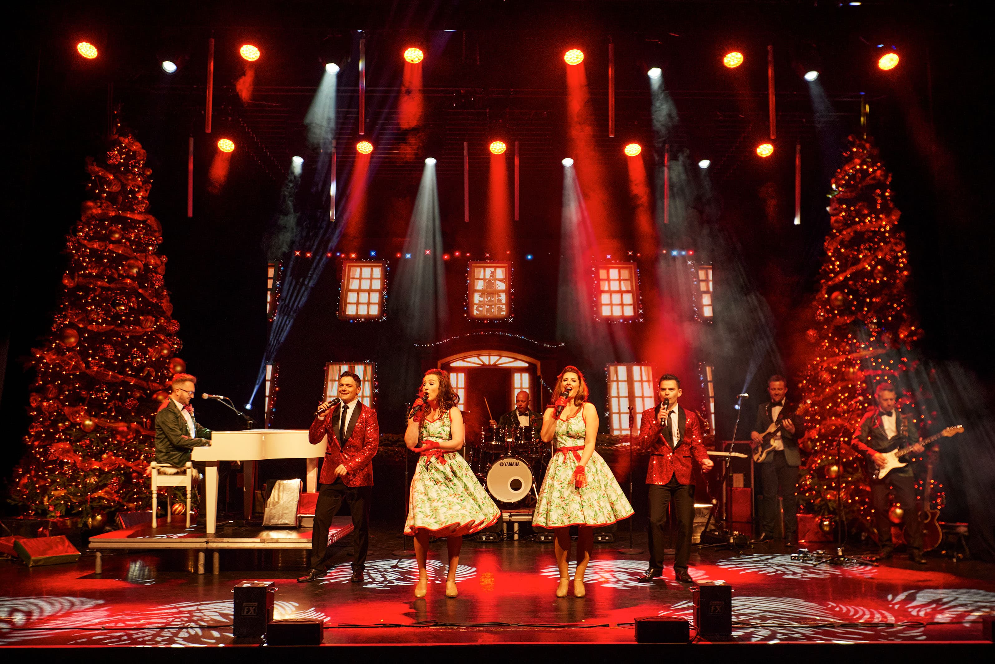 A group of musicians and singers perform on stage, surrounded by large decorated Christmas trees, celebrating the festive season.